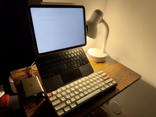 A tiny desk in a dimly-lit corner. There's an iPad, a mechanical keyboard, and a portable DAC. - [Header image for the post: My Setups Part One - Writing This - Here, I talk about one of my setups (part one)]